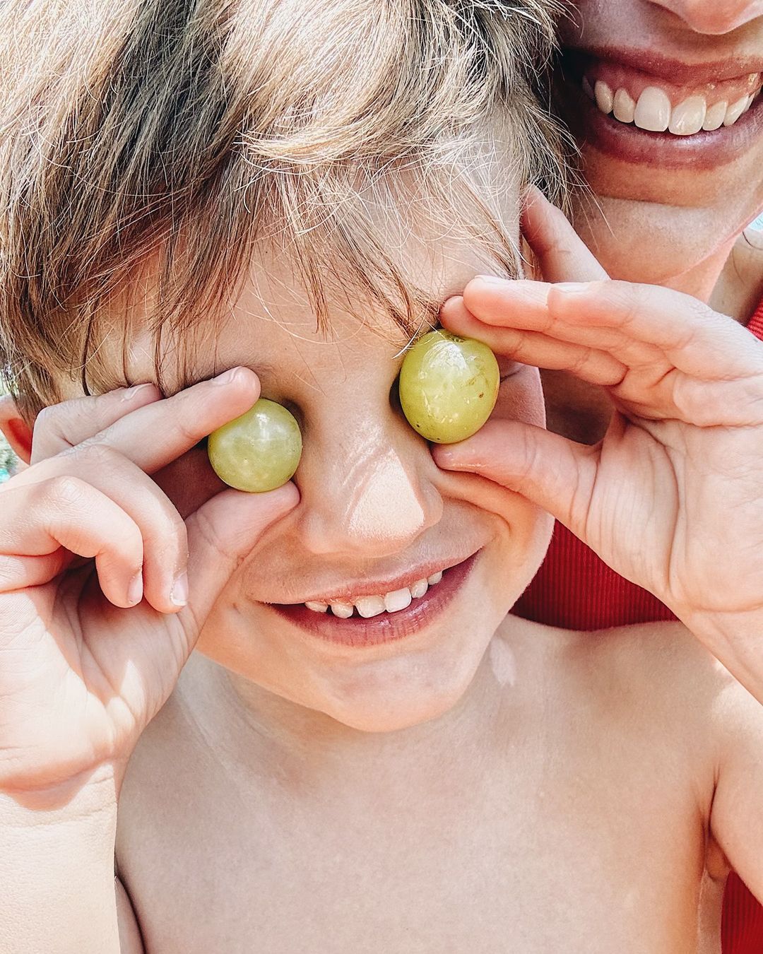 Happy child holds grapes up to face like eyes, grinning at the camera with an adult in the background; summer, daylight.