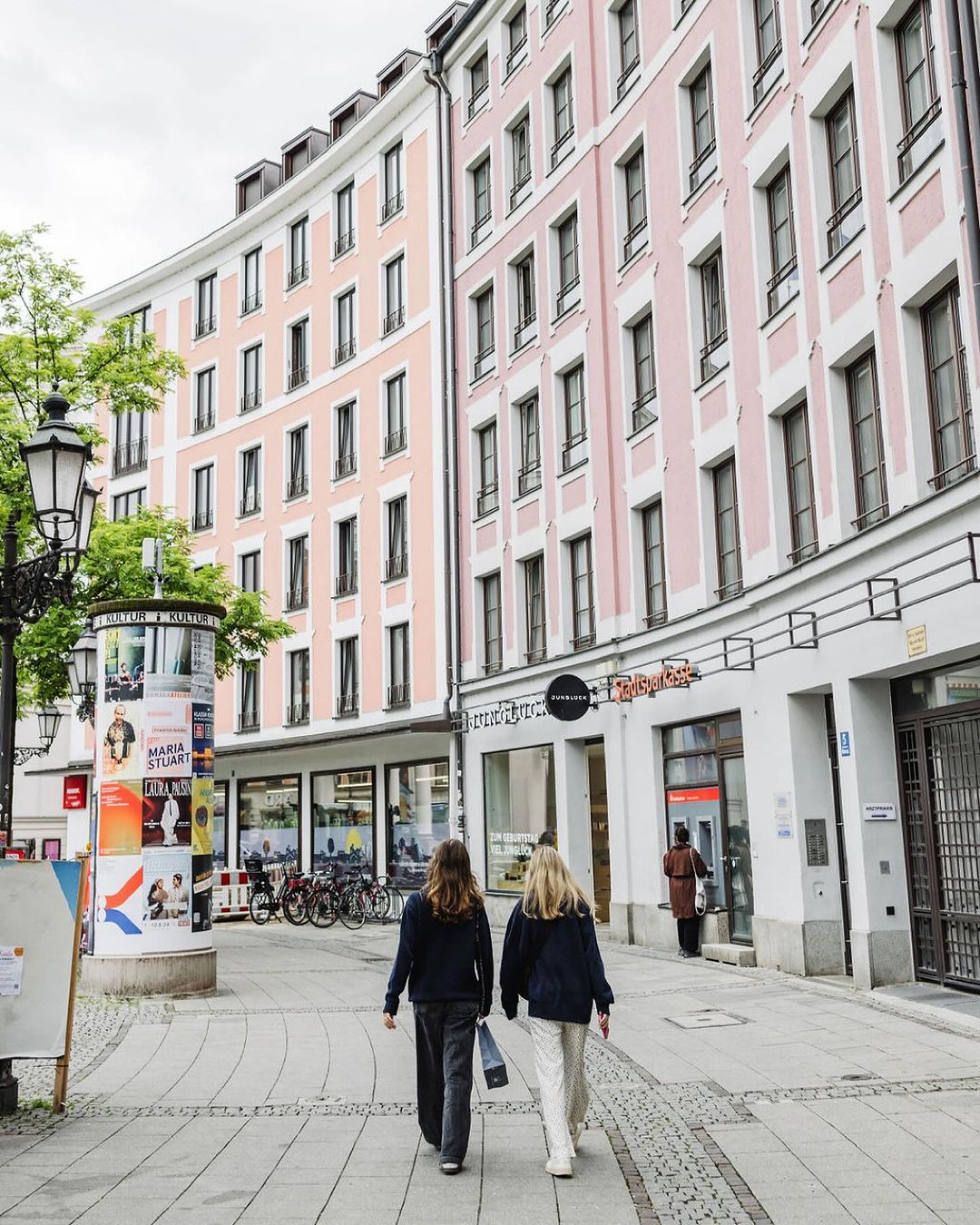 Two adult females walk along a cobblestone street lined with pink buildings, shops, trees and a lamp post with posters in an urban area.