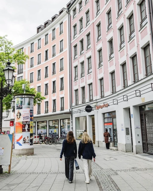 Two adult females walk along a cobblestone street lined with pink buildings, shops, trees and a lamp post with posters in an urban area.