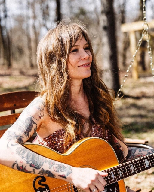 Woman with long brown hair and tattoos plays an acoustic guitar outdoors, seated on a wooden bench near trees and a swing.