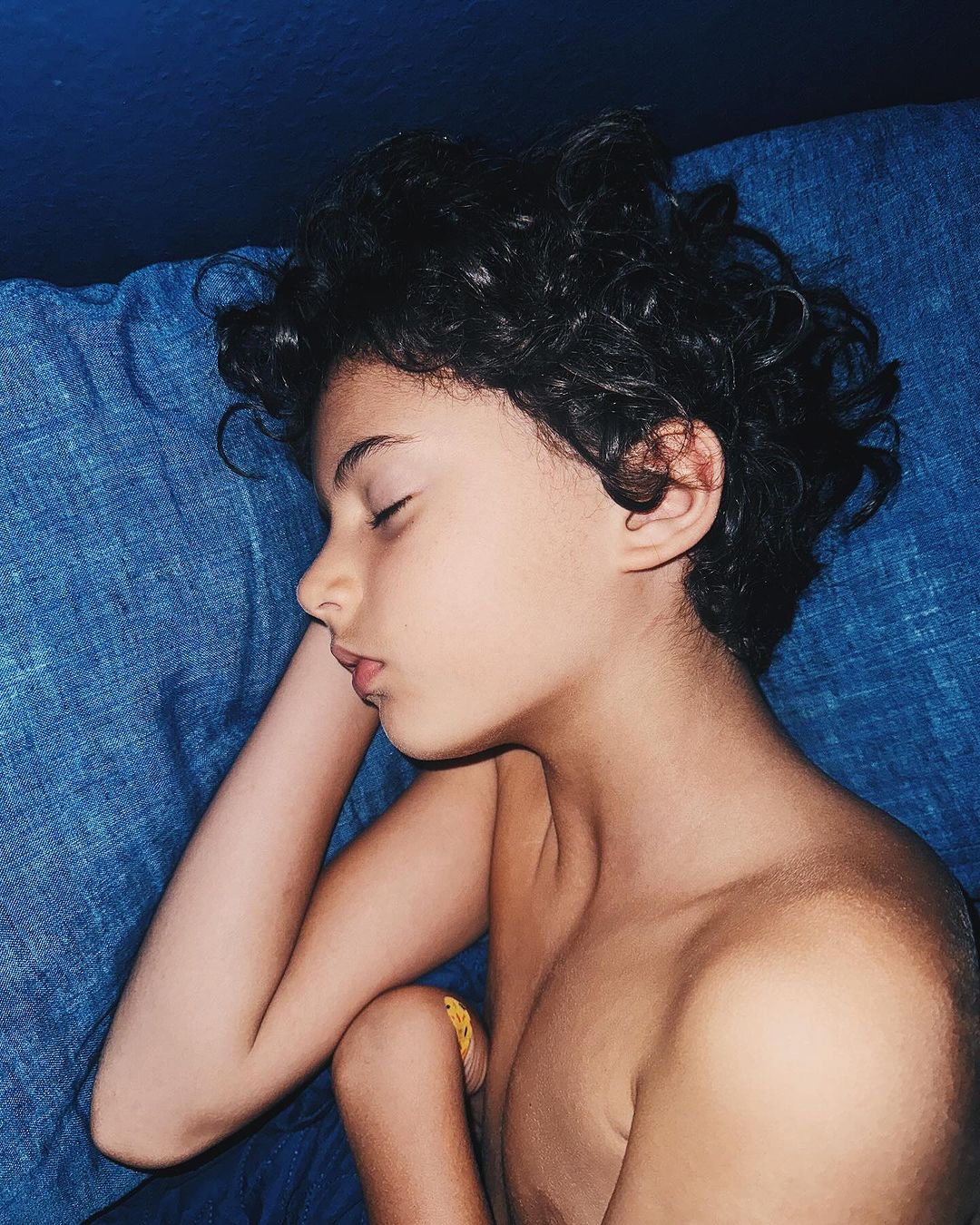 Close-up portrait of a young child with dark curly hair sleeping soundly against a blue textured pillow.