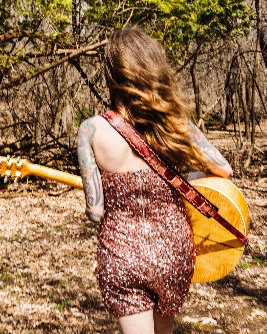 Back view of a musician in a sparkling dress walking through a sunny forest with a banjo, creating a harmonious blend of fashion and nature.