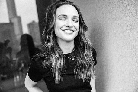 Monochrome portrait of a smiling woman with long wavy hair wearing a t-shirt and necklace, photographed by Stephanie Bassos.