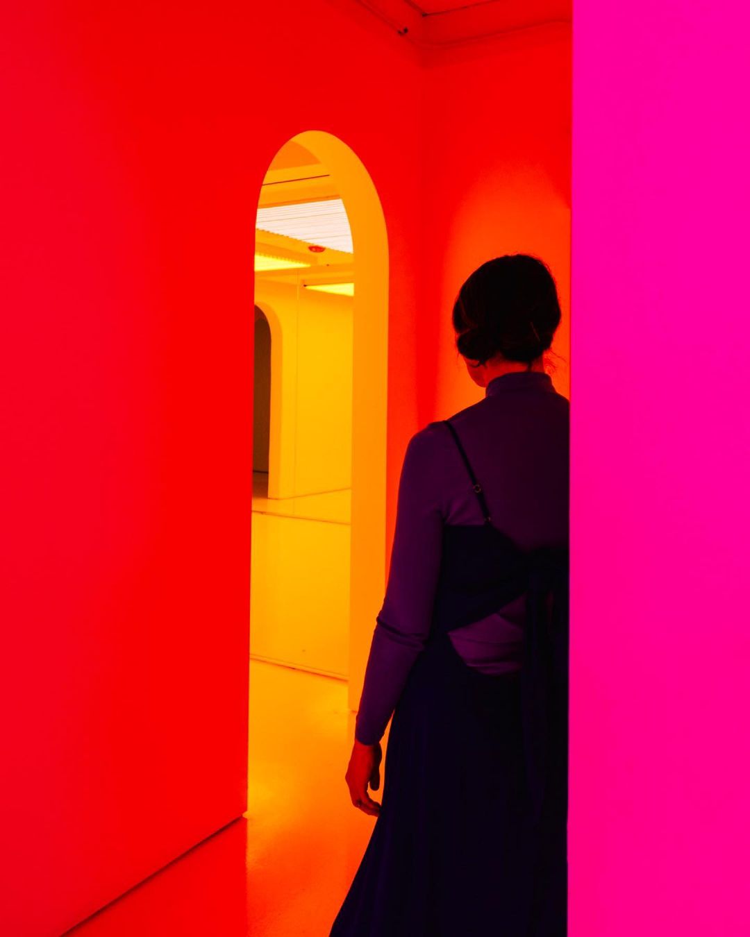Woman in dark dress walks through brightly lit archway with red and pink walls. Interior design features contrasting saturated colors.