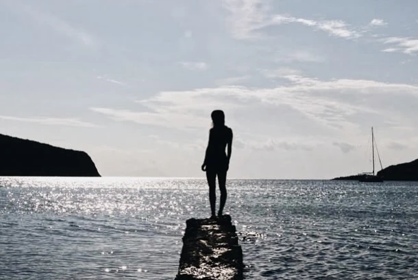 Silhouette of a woman standing on a rock jetty in the ocean, with a sailboat visible in the distance, taken during daylight.