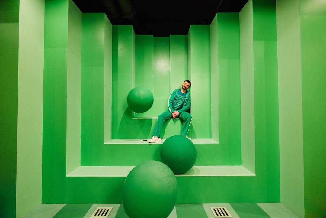 Man in green tracksuit leans against matching wall with large green spheres in an indoor studio-like setting with striped floor.
