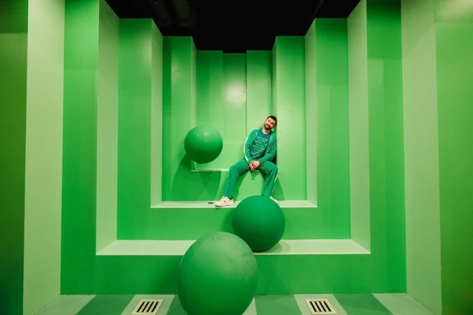 Man in green tracksuit leans against matching wall with large green spheres in an indoor studio-like setting with striped floor.