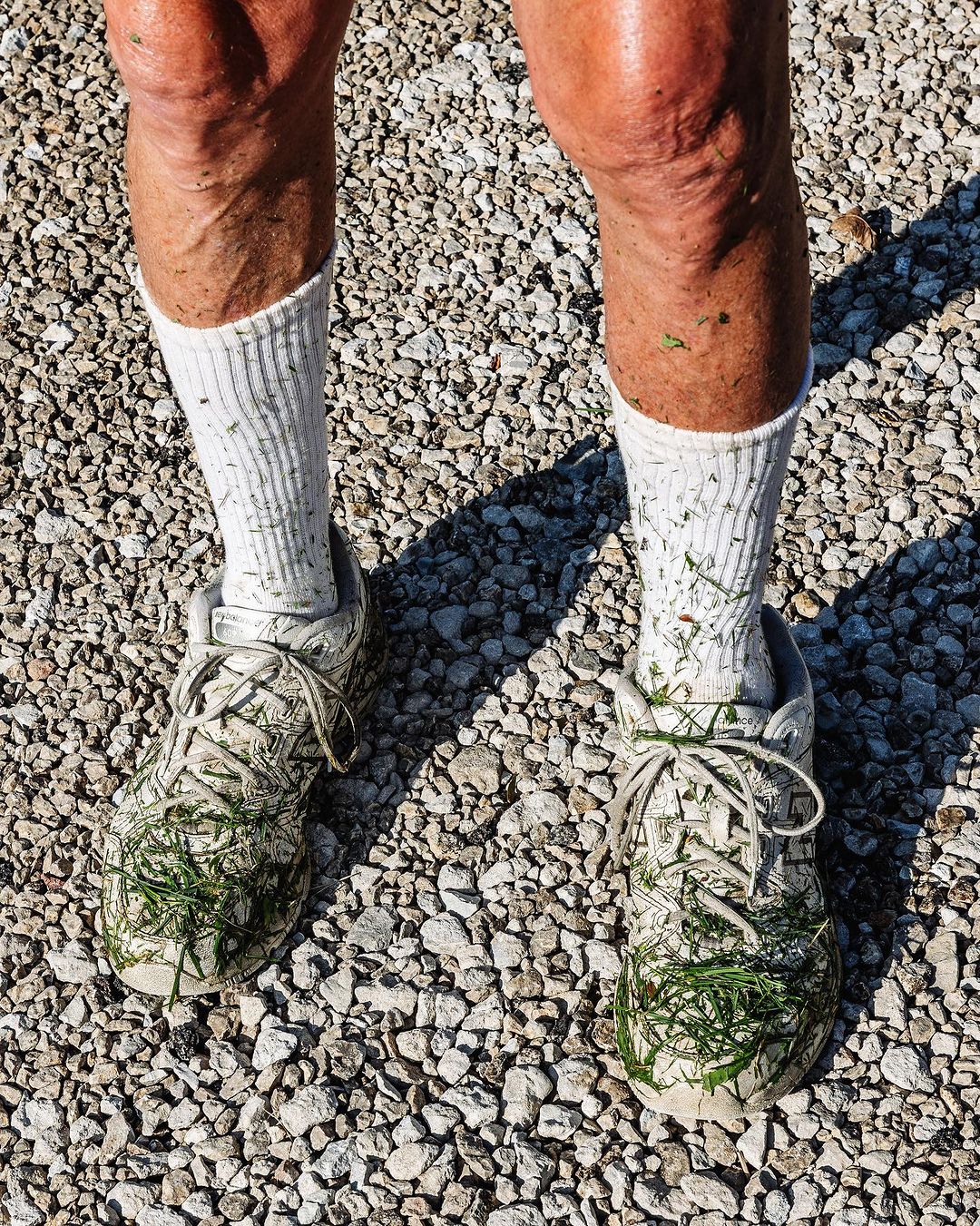 Close-up of an adult's legs and feet wearing white sneakers covered in grass clippings, standing on a gravel surface in bright sunlight.