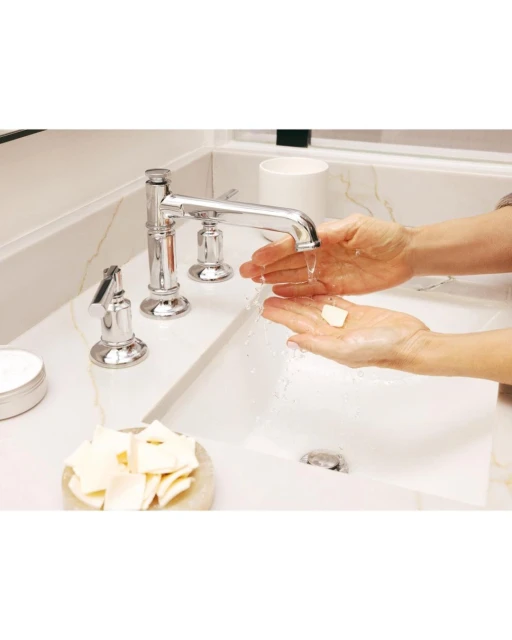 Woman washing hands at marble sink with running chrome faucet and white cleansing squares, in a light-filled bathroom setting.