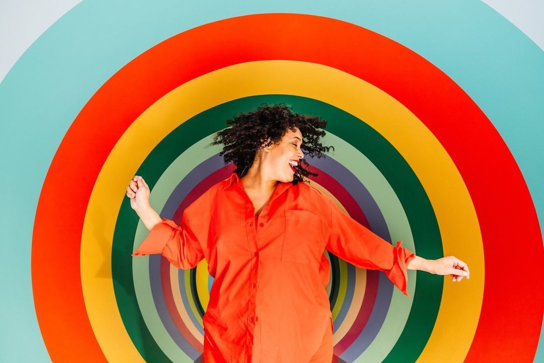 Smiling woman with curly hair in an orange shirt poses in front of a colorful circular rainbow background.