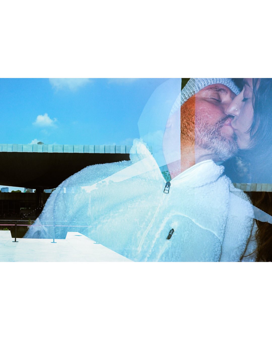 Double exposure of a man and woman kissing, set against a blue sky with a bridge, blending portrait and urban landscape elements.