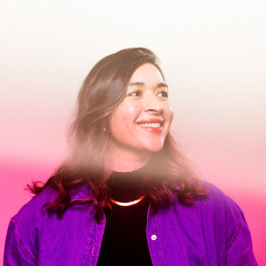 Portrait of a smiling woman with long brown hair wearing a purple jacket and gold necklace against a vibrant pink and white background.