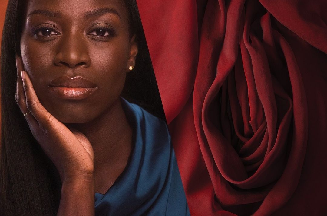 Portrait of a dark-skinned woman with hand on her face beside a red fabric shaped like a vulva. High contrast, striking image.