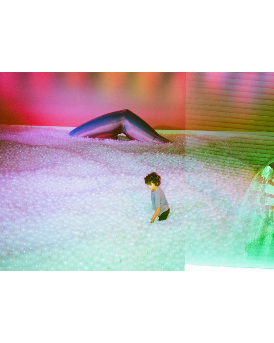 A child with curly hair sits in a large pit filled with white balls. A blue archway is visible in the background, illuminated by pink light.