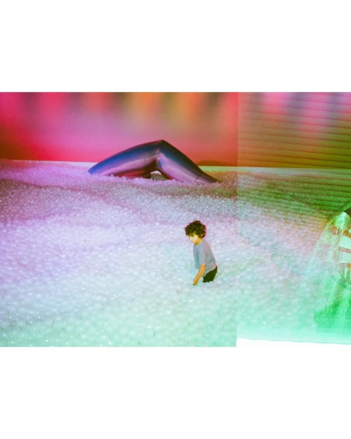 A child with curly hair sits in a large pit filled with white balls. A blue archway is visible in the background, illuminated by pink light.