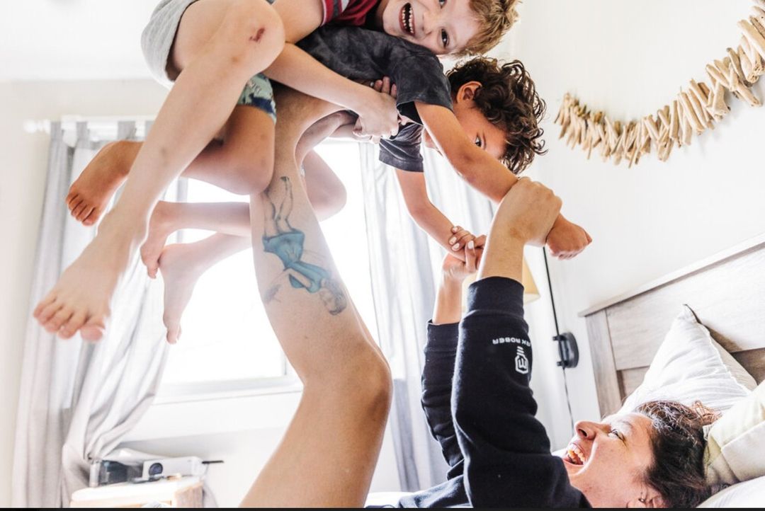 A happy family moment indoors: a dad lying on a bed lifts two smiling young children in the air, creating a joyful, playful scene.