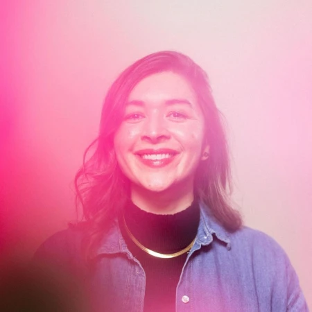 Portrait of smiling woman in black turtleneck and denim shirt bathed in pink light by Stephanie Bassos