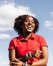 Smiling black woman wearing a red polo shirt with beaded bracelets against a clear blue sky with fluffy white clouds.