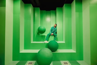 Man in green tracksuit seated in a stepped green alcove with three green spheres by Stephanie Bassos