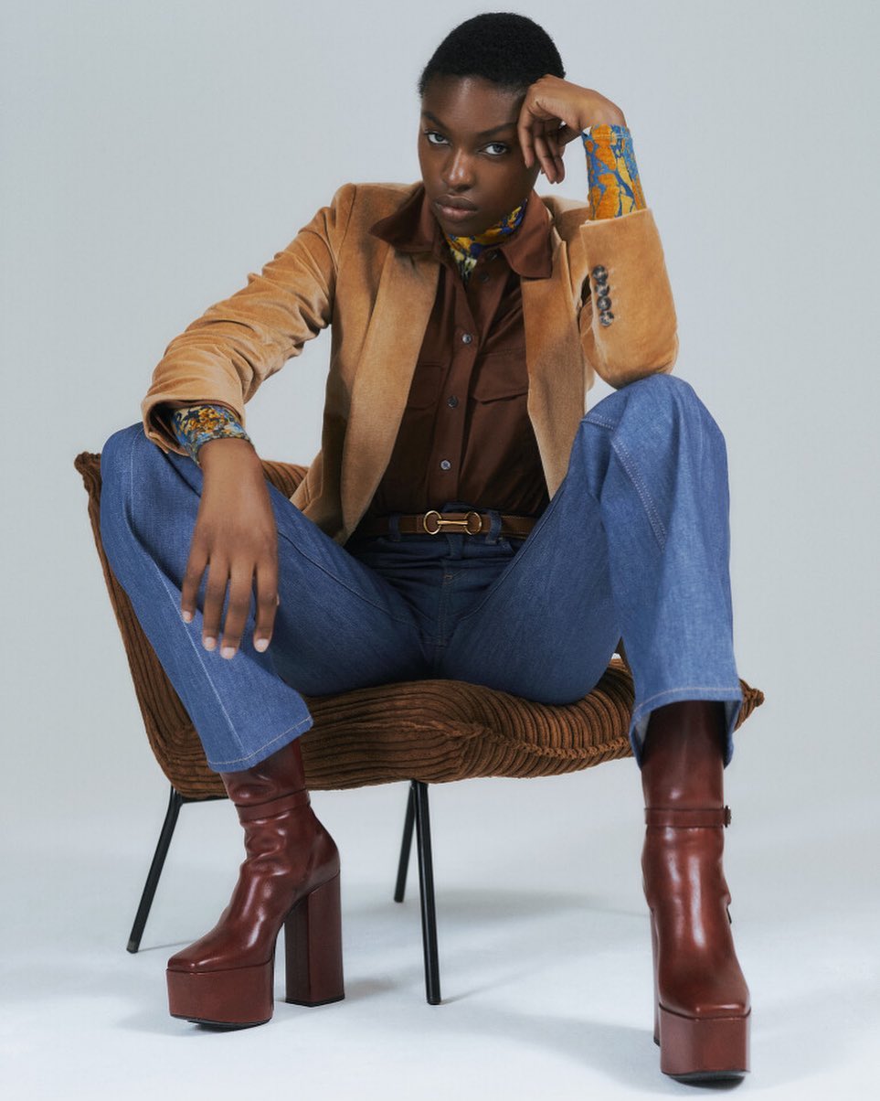 Elegant African-American woman in a tan blazer, brown shirt, and blue jeans seated in a chair, highlighted by studio lighting.
