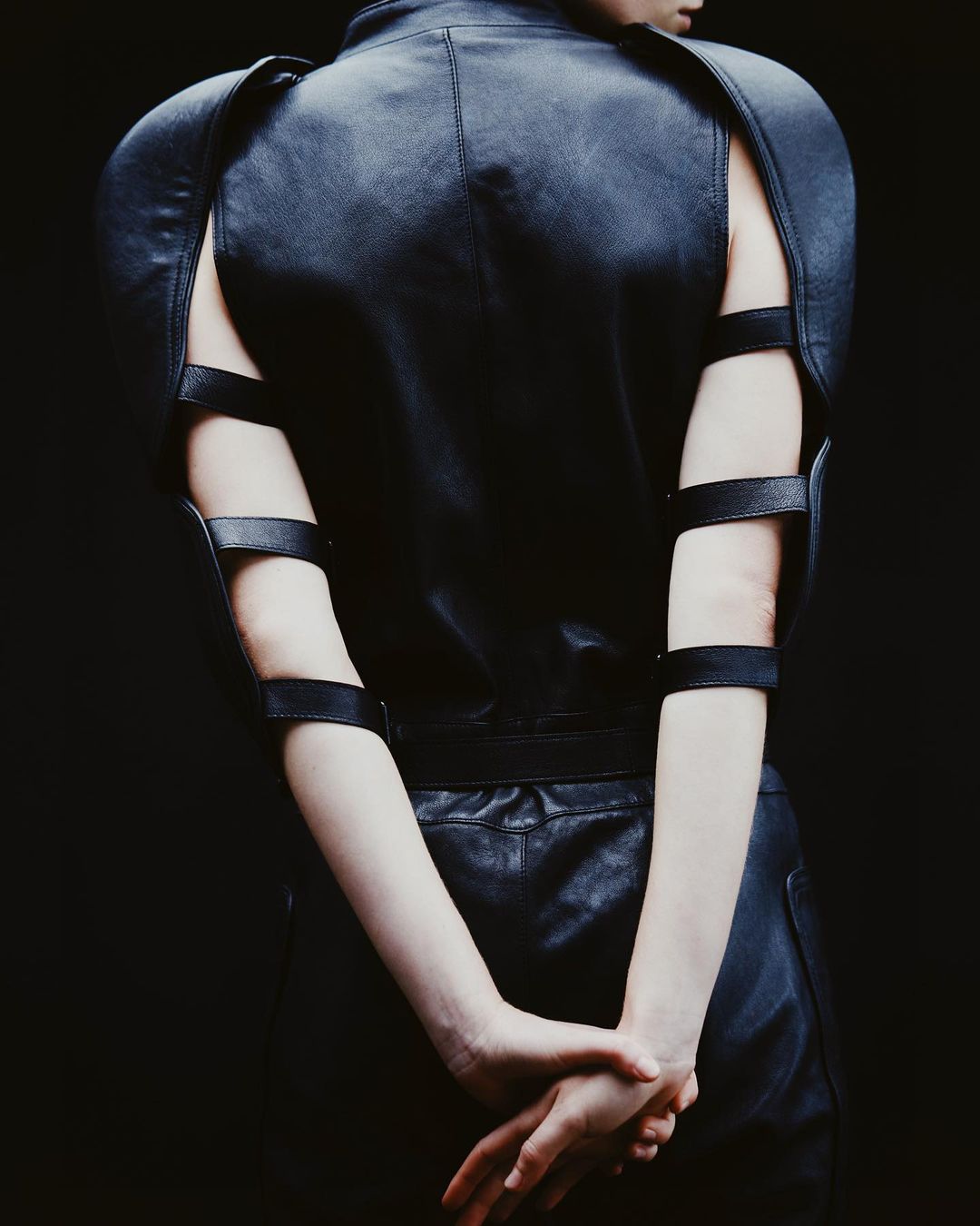 Back view of a slender woman in a dramatic black leather outfit with angular shoulders and arm straps, hands clasped in a dark studio.