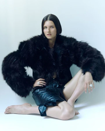 Woman in a black faux fur jacket and dark leather shorts sitting against a seamless white studio background