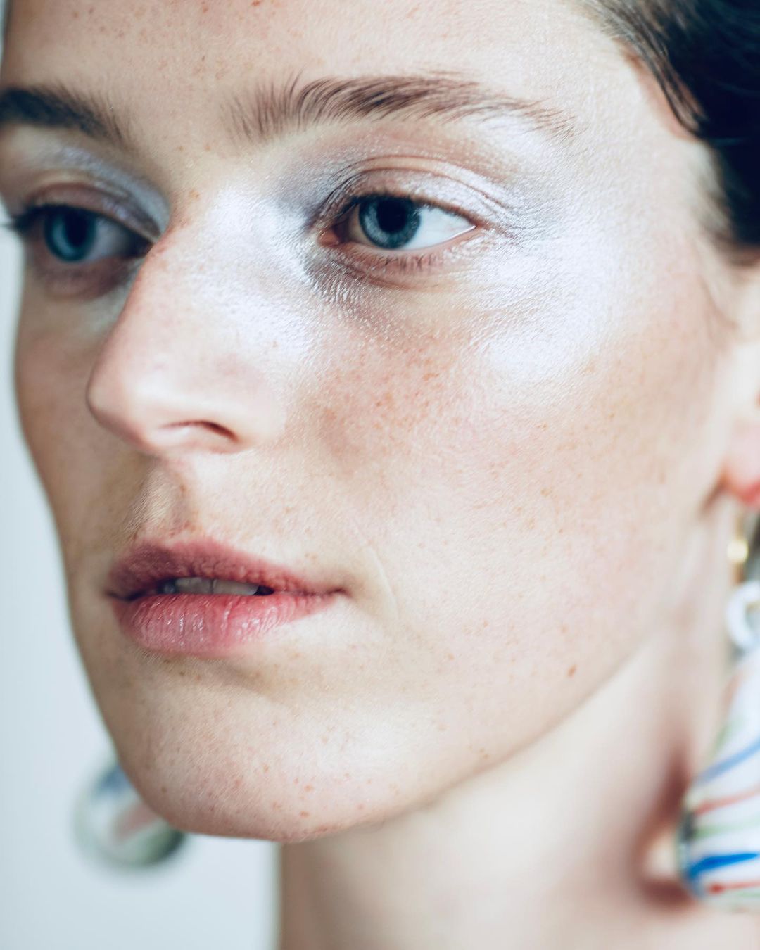 Close-up portrait of a woman with blue eyes and glowing silver eyeshadow. She wears statement earrings and has fair skin with freckles.