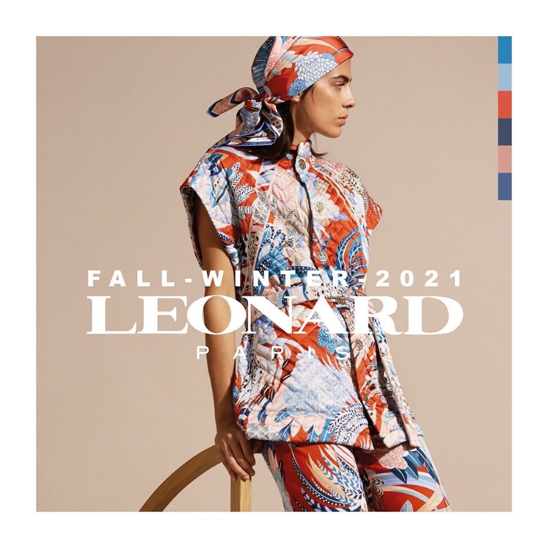 Fashion model wearing a coordinated floral print headscarf, quilted vest, and pants poses near a wood accent chair in a studio.