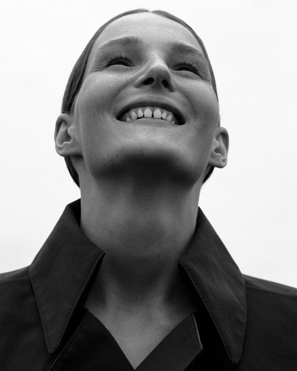Black and white portrait of a smiling adult woman looking upwards, wearing a coat, against a neutral sky background.
