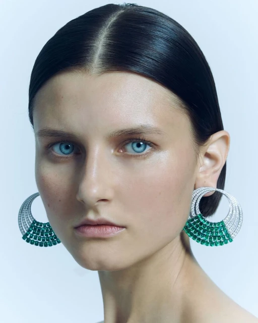 Close-up of a woman with light skin, blue eyes, and dark hair, wearing large, circular earrings with white and green gemstones.
