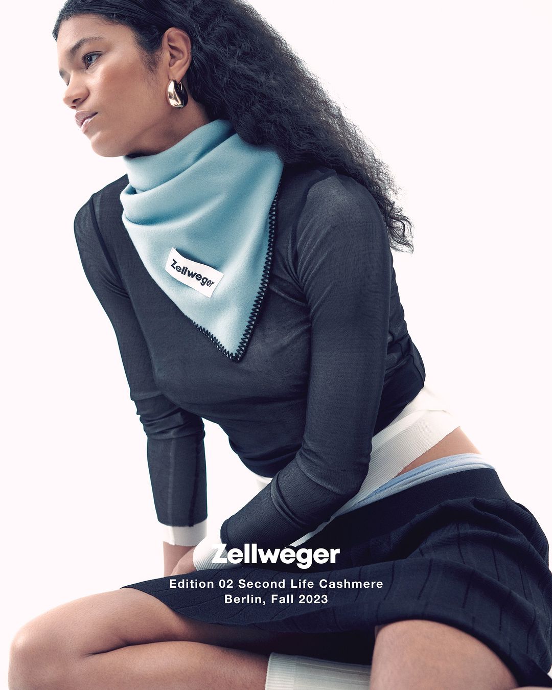 Young woman in a dark long-sleeve shirt and skirt poses with a light blue scarf against a bright background in a fashion setting.