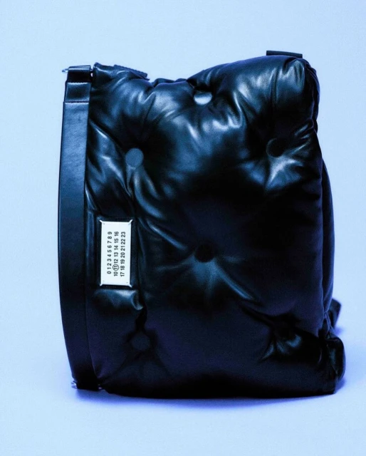 Close-up of a black quilted puffy bag with a white tag displaying numbers and a black strap, displayed against a blue background.