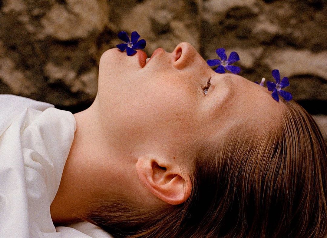Close-up studio shot of a light-skinned woman lying down with dark violet flowers adorning her face, wearing a white top against a stone wall.