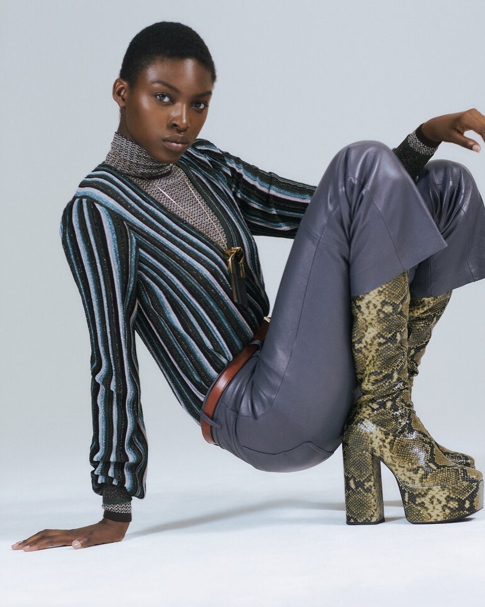 A Black female fashion model poses in a studio wearing a striped top, grey leather pants, and snake print boots in a fashion editorial.