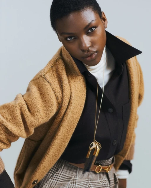 Fashion shot of a Black woman in a brown cardigan and plaid pants, wearing a black button-down shirt and gold necklace against grey backdrop.