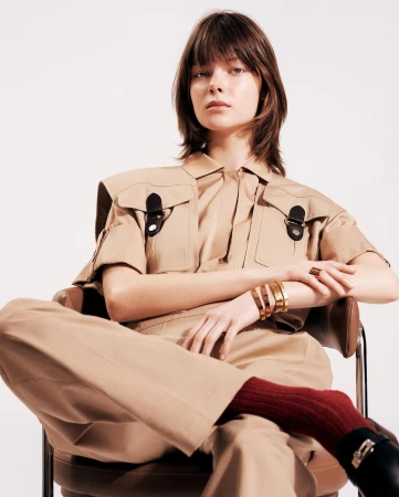 Model seated in a chair wearing a tan utilitarian jumpsuit, gold bangles, and dark red socks photographed by Stephanie Pfaender.