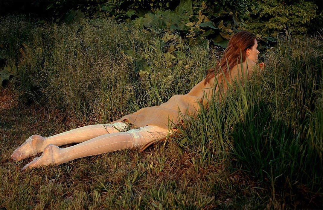 Adult female with long brown hair poses while lying in a grassy field wearing sheer stockings in an outdoor environment.