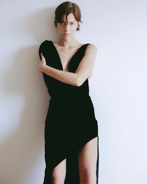 A woman in a black dress poses in a studio with arms crossed, short brown hair, fair skin, indoors with white wall background.