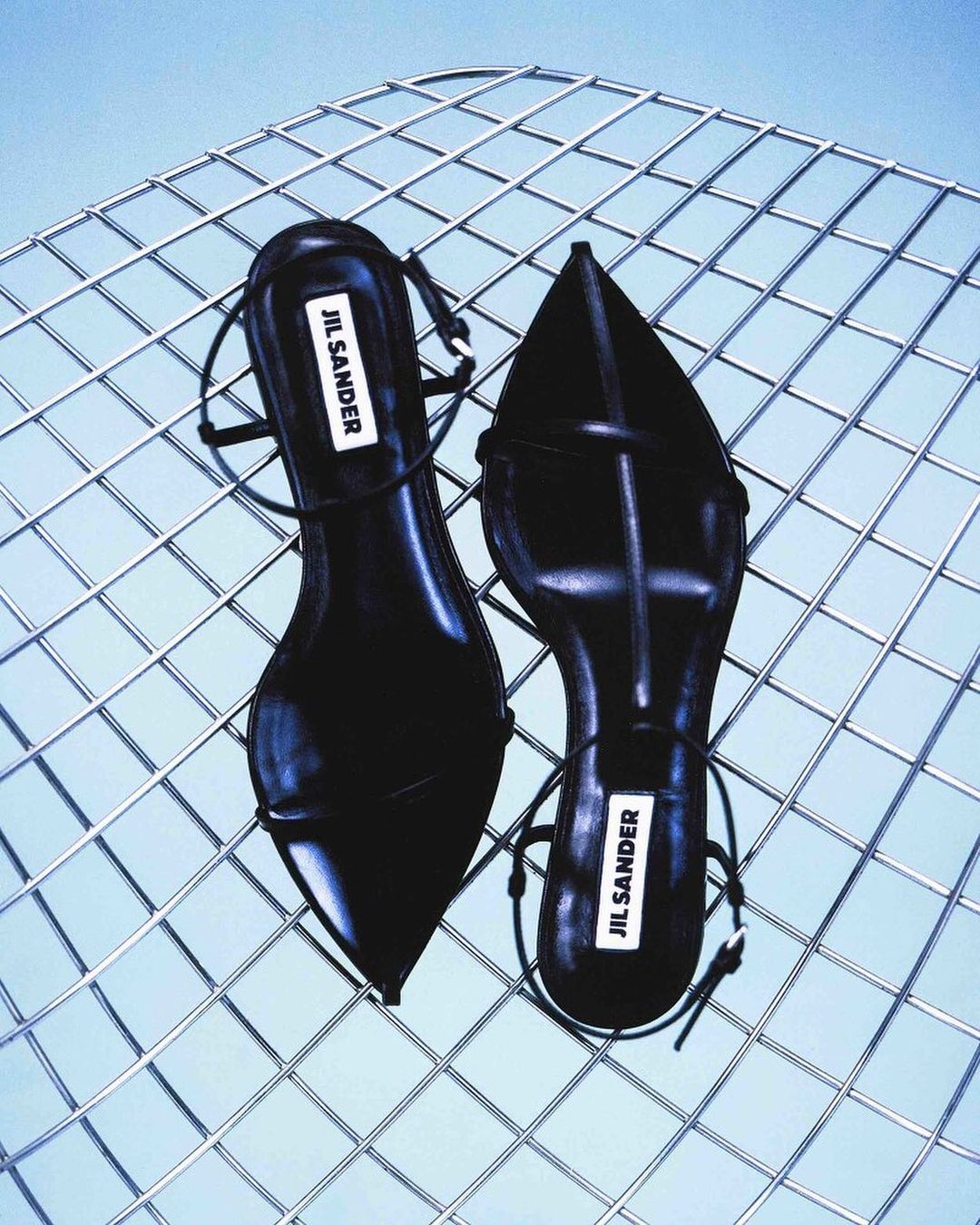 Overhead shot of Jil Sander black leather heeled sandals displayed on a silver metal wire grid with a light blue background.