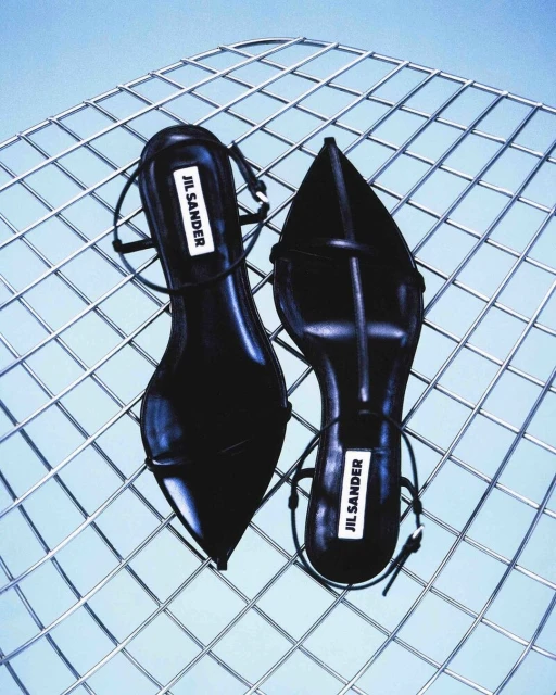 Overhead shot of Jil Sander black leather heeled sandals displayed on a silver metal wire grid with a light blue background.