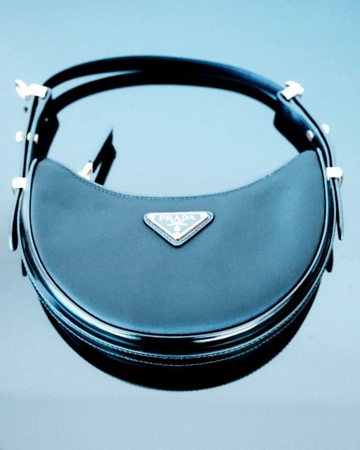 Black Prada Cleo handbag on a light blue surface. Features a semi-circular shape, leather trim, metal hardware, and a triangular logo.