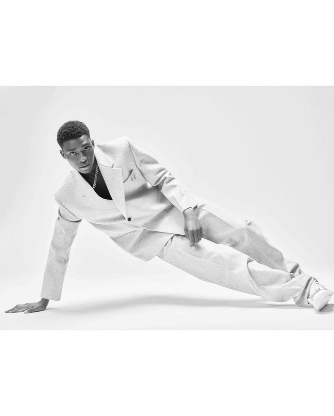 Monochrome portrait of a stylish young Black man in a suit striking a dynamic pose against a plain white background in a studio setting.
