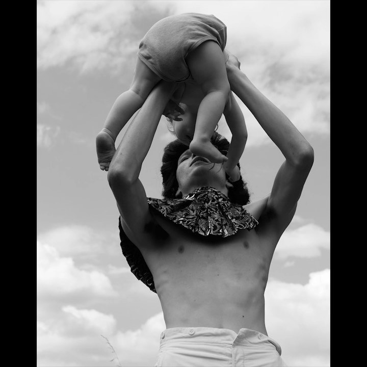 Black and white photo of an adult holding a baby overhead, with a cloudy sky in the background. The adult is shirtless with patterned fabric around their neck.