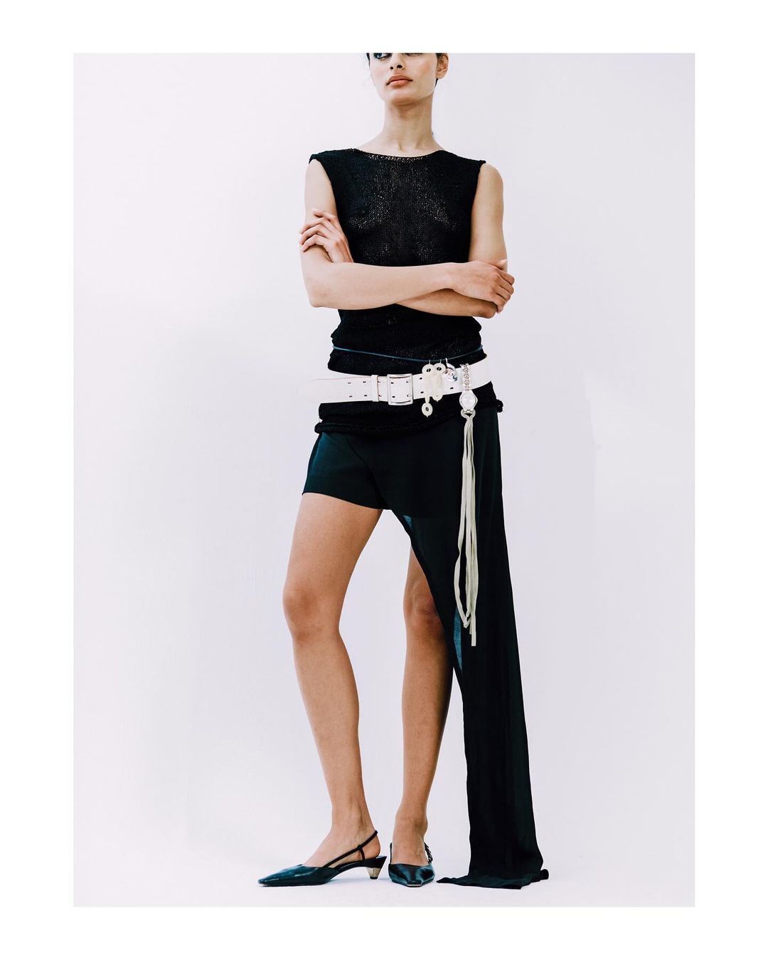 A stylish adult female stands in a photo studio wearing a black sleeveless top, a white belt, black shorts, and black slingback heels.