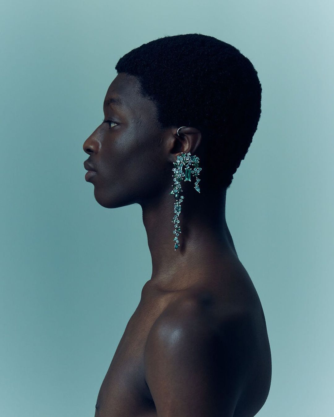 Side profile studio shot of a shirtless African-descent male model with large, elaborate dangling earring in a blue-toned studio environment.