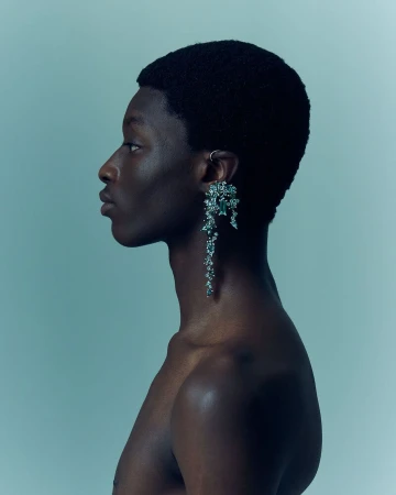 Side profile studio shot of a shirtless African-descent male model with large, elaborate dangling earring in a blue-toned studio environment.