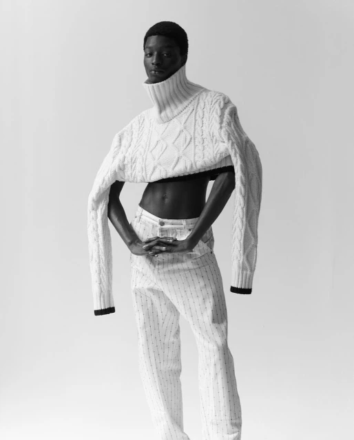 Black model poses in cropped cable-knit sweater and striped pants against a white backdrop in a studio portrait, black and white.