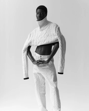 Black model poses in cropped cable-knit sweater and striped pants against a white backdrop in a studio portrait, black and white.