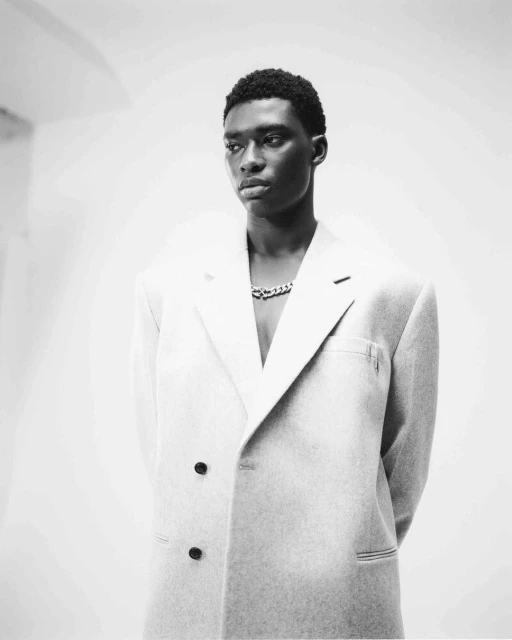 Black and white portrait of a Black male model in a light-colored suit jacket and a chain necklace against a plain background.
