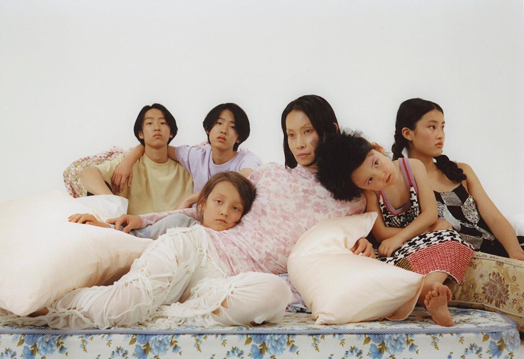 Diverse group portrait features an Asian adult female and four children of Asian descent in fashionable clothing, posed on a floral mattress.